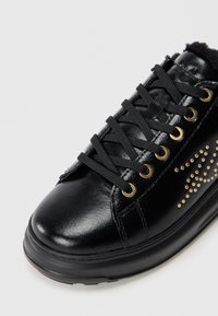 Svart lær sneaker med gulløyer, svarte snorer og en teksturert overflate med naglede detaljer på siden. Flat gummisåle.