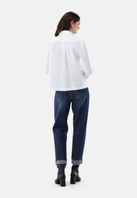 Blusa bianca a maniche lunghe con piega sul retro, abbinata a jeans larghi blu scuro e stivali neri con tacco, che mostrano linee pulite e un design semplice.