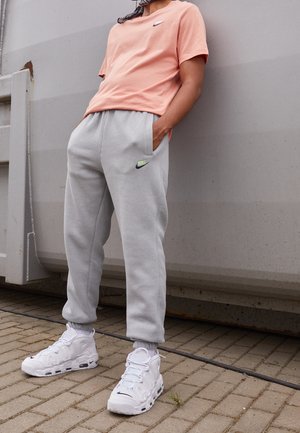 Person trägt pfirsichfarbenes Nike T-Shirt, hellgraue Nike Jogginghose und weiße Nike Sneakers, steht vor einer grauen Metallwand auf gepflastertem Boden.