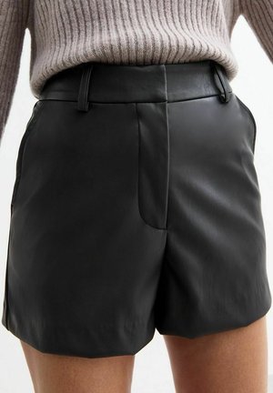 Close-up van een persoon die zwarte leren shorts en een beige geribde trui draagt, ingestopt, met de tailleband en voornaad details zichtbaar.