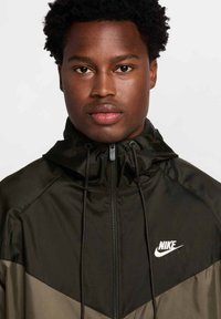 Kurtka z kapturem Nike wykonana z gładkiego czarnego i oliwkowego materiału, z zamkiem błyskawicznym z przodu, sznurkiem ściągającym oraz białym logo na klatce piersiowej.