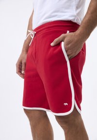 Czerwone spodenki sportowe z białym wykończeniem, elastycznym pasem i sznurkiem do regulacji. Posiadają kieszenie oraz małe logo z boku. Wykonane z miękkiego materiału bawełnianego.