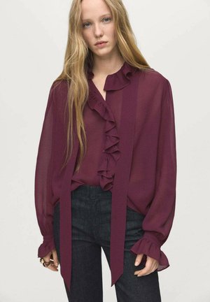 Blouse en voile bourgogne avec un devant à volants, manches longues avec des poignets à volants, et de longs liens à la taille. Portée avec un pantalon en denim foncé.