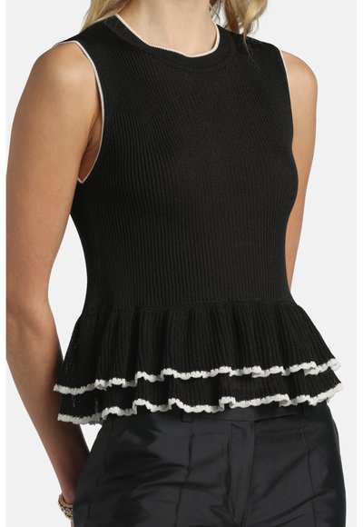 Haut noir sans manches à côtes avec un ourlet à volants double couche bordé de blanc, porté avec un pantalon noir et des bracelets dorés au poignet gauche.