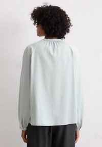 Jasnoniebieska bluzka z długim rękawem, uszyta z gładkiego materiału, z dekoltem w falbanki i guzikami przy mankietach, noszona z ciemnymi spodniami.