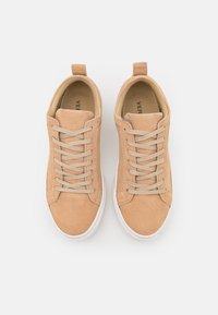 Sneakers de camurça em tom beje com uma textura suave, atacadores planos e sola de borracha branca. Apresenta uma alça na parte traseira para facilitar o calçar.