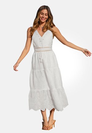 Kobieta uśmiechająca się, z długimi falującymi włosami, ubrana w białą sukienkę midi z ażurowym wzorem bez rękawów oraz beżowe sandały na wysokim obcasie, z wyciągniętymi ramionami.