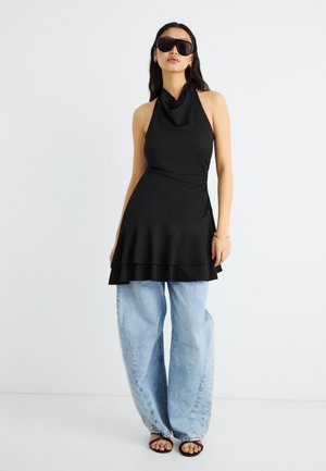 Kvinde iført stor sort haltertop, lyseblå vide jeans, sorte sandaler, store mørke solbriller, stående mod en hvid baggrund.