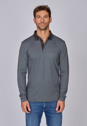 Homme se tenant face à l'avant, portant un polo gris à manches longues et un jean bleu, sur un fond gris clair uni.