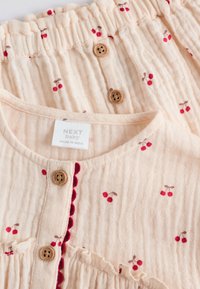 Tenue en mousseline couleur pêche avec motifs de cerises rouges, boutons en bois, accents froufroutants et bordure festonnée sur le col.