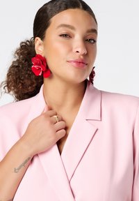 Roze blazer met revers, gedragen over een witte top. Grote rode bloemvormige oorbellen en een gouden ring zijn opvallende accenten. Glad textuur duidelijk zichtbaar.