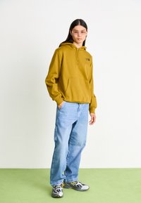 Olive green hooded sweatshirt with front pocket, worn with light blue baggy jeans and black sneakers featuring yellow accents. Plain backdrop.