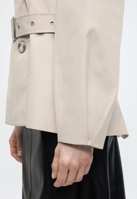 Μανίκι μπεζ καμπαρντίνας με λεπτομέρειες κουμπιού και ζώνης, φορεμένο πάνω από μαύρη πλισέ φούστα, με ένα χαλαρό χέρι στο πλάι.