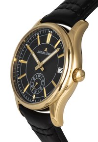 Guldtonad klocka med svart urtavla, guldmarkörer för timmar, subdial och datumsdisplay. Har ett svart läderarmband och en rund klockkapsel.