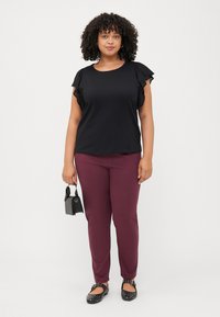 A woman stands in a black top with ruffled sleeves and maroon pants, holding a small black handbag. She wears black studded flats.