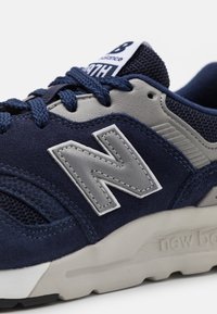 Marinblå atletisk sneaker gjord av mocka och nätmaterial, med en grå "N"-logotyp, texturerade detaljer och en l ätt beige sula.