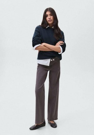 Femme debout, les bras croisés, portant un pull marine par-dessus une chemise blanche, un pantalon large marron et des ballerines noires à clous.
