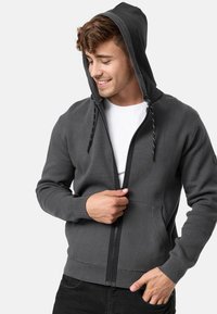 Sweat à capuche zippé gris avec cordon de serrage, poignets côtelés et poches avant. Fabriqué en tissu doux avec une texture lisse.