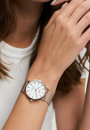 Frau trägt eine Armbanduhr mit einem roségoldenen Mesh-Armband, weißem rundem Zifferblatt, goldenen Stundenmarkierungen und drei Zeigern, die auf 6:25 Uhr eingestellt ist.