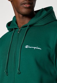 Groene hoodie gemaakt van zacht materiaal, met een rits aan de voorkant, koordjes en een wit "Champion" logo op de borst.