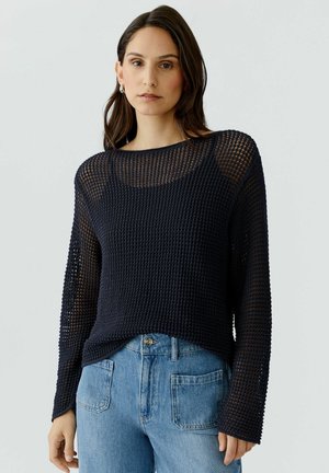Femme portant un haut noir à manches longues en maille ajourée par-dessus un débardeur noir et un jean taille haute bleu clair, debout devant un fond uni.