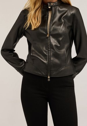 Femme portant une veste en cuir noire ajustée avec une fermeture éclair devant et un col montant, associée à un pantalon noir, les mains derrière le dos.