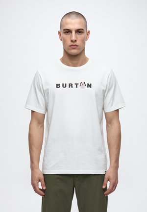 Un homme porte un T-shirt blanc Burton orné d’un petit logo et d’un motif graphique, associé à un pantalon vert olive, sur un fond uni et clair.