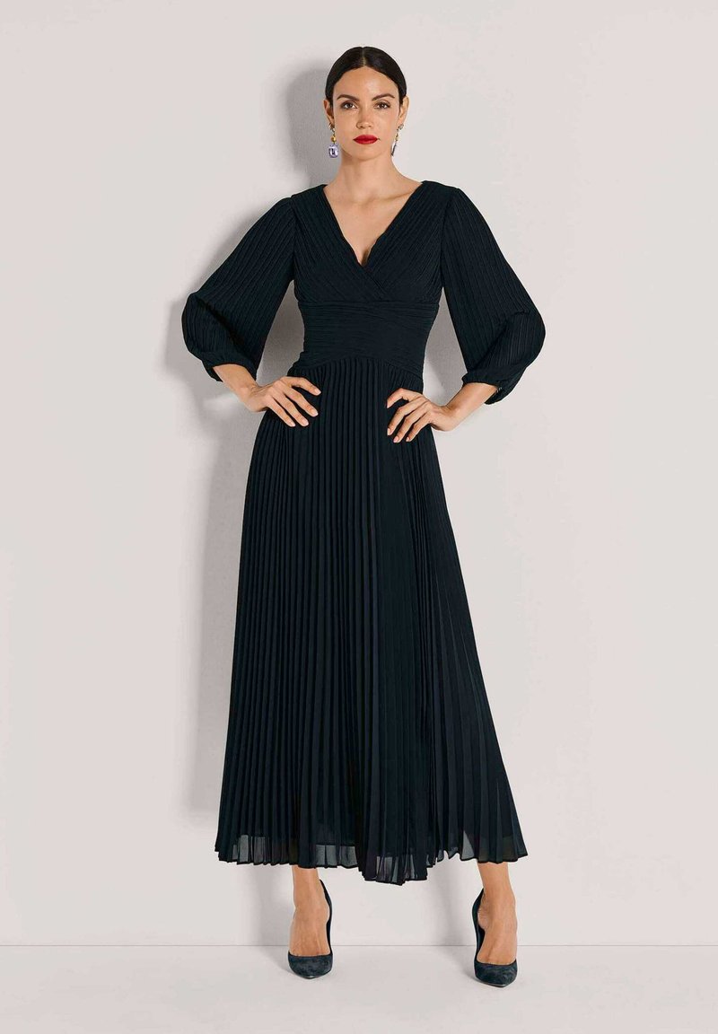 Zwarte geplooide jurk met een V-hals, lange pofmouwen en een nauwsluitend lijfje, uitlopend in een vloeiende, enkelhoge rok.