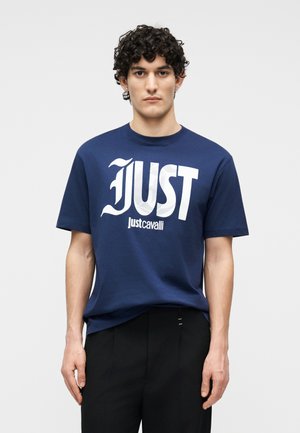 Mężczyzna z kręconymi włosami, ubrany w granatowy T-shirt Just Cavalli i czarne spodnie, stojący na tle gładkiego szarego tła.