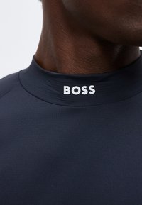 Κοντινό πλάνο ενός ατόμου που φοράει σκούρο μπλε μπλουζάκι με ψηλό γιακά με τη λέξη «BOSS» με λευκά γράμματα στο γιακά.