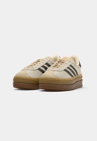 Beige and tan suede sneakers with a thick brown sole, featuring three blue stripes and cream laces. Rounded toe design, minimal accents.
