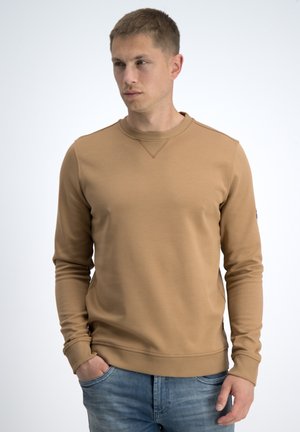 Hombre con sudadera de cuello redondo color tostado y vaqueros azules, de pie con una mano en el bolsillo, mirando de lado contra un fondo blanco liso.