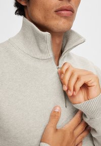 Svijetlosiva, zip-up dukserica s rebrastim ovratnikom i teksturiranim materijalom. Ruke drže zip blizu vrata. Bliski pregled.