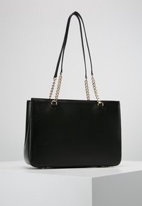 Sac cabas noir avec une finition texturée brillante, double bandoulière en cuir noir et accents de chaîne dorée, présentant une forme rectangulaire structurée.