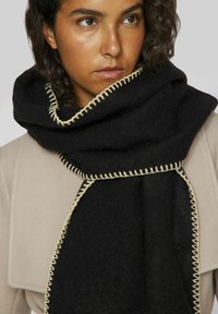 Bufanda de lana negra con una textura suave, con un contraste de costura en beige claro a lo largo de los bordes, envuelta alrededor del cuello del modelo.