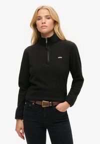 Zwarte fleecetrui met een hoge kraag en een halflange ritssluiting; heeft een klein logo op de borst en lange mouwen. Gecombineerd met donkere jeans.