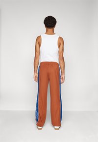 Bruine wijde broek met blauwe zijkanten en witte logo-details, gecombineerd met een witte mouwloze top, gezien van achteren.