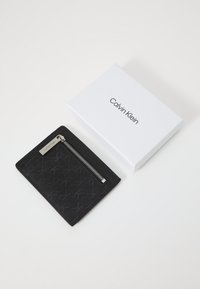 Portefeuille noir à texture avec une poche zippée. Présente un motif subtil et un accent en métal argenté. Livré dans une boîte blanche.