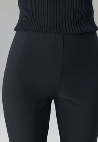 Schwarze, hoch taillierte Hosen aus Stretchstoff mit glatter Textur und dezenten Seitentaschen. Oberteil mit Rippstruktur, das über dem Hosenbund sichtbar ist.