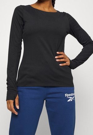 Femme portant un t-shirt noir moulant à manches longues et un pantalon de jogging bleu Reebok, avec une main sur la hanche et l’autre détendue.
