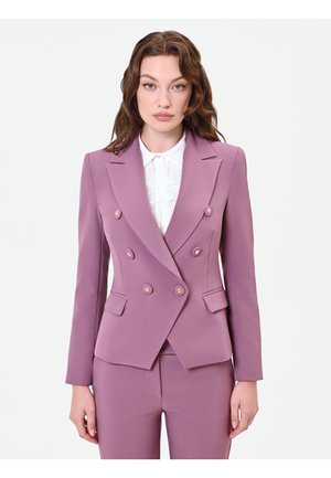 Donna con capelli castani ondulati indossa un blazer doppiopetto color malva su misura con bottoni sopra una camicetta bianca con colletto di pizzo e pantaloni coordinati.