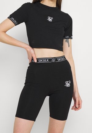 Kobieta nosząca czarny krótki top i wysokie spodenki rowerowe z białymi logo SikSilk na piersiach, rękawach, pasku i udzie.