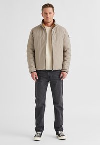 Helle beige gesteppte Jacke mit hohem Kragen, schwarzer Reißverschluss und orangefarbener Futter, getragen über einem cremefarbenen Pullover und kombiniert mit dunklen Jeans.