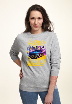 Kvinde iført grå sweatshirt med farverig grafik af en blå bil, graffiti tekst og frasen "New Standard" i gule og lilla toner.