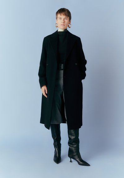 Svart ullrock med dubbelknäppt design, kombinerad med en åtsittande svart polotröja och höga boots. Har stora guldörhängen i form av ringar.