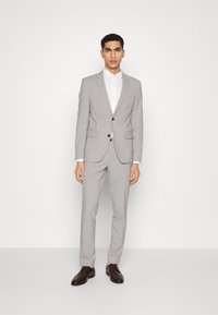 Completo grigio chiaro con vestibilità slim, giacca monopetto, bottoni neri e pantaloni coordinati. Camicia bianca sotto, abbinata a scarpe marroni scure.