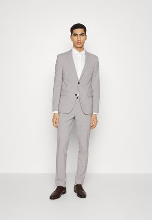 Costume gris clair avec une coupe slim, veste à un bouton, boutons noirs et pantalon assorti. Chemise blanche en dessous, accompagnée de chaussures marron foncé.