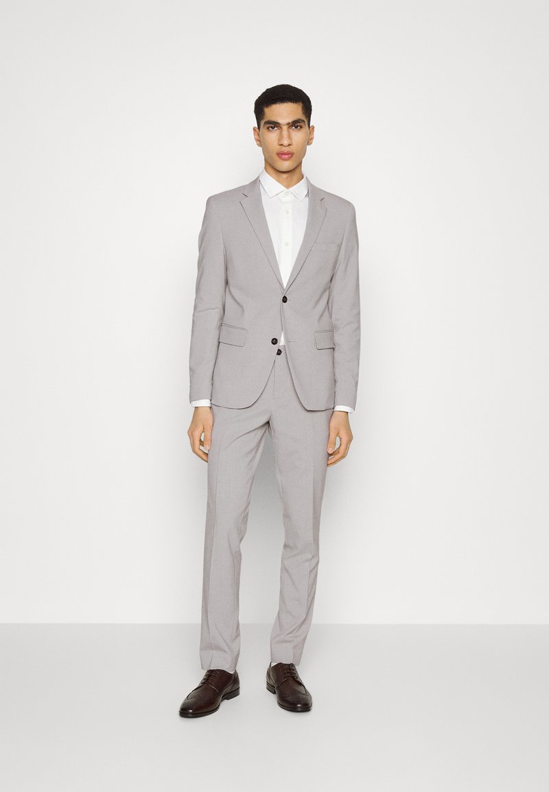 Completo grigio chiaro con vestibilità slim, giacca monopetto, bottoni neri e pantaloni coordinati. Camicia bianca sotto, abbinata a scarpe marroni scure.