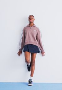 Rudas zip-up džemperis su baltais dryžiais, derinamas su tamsiu mini sijonu. Juodi batai su purpuriniais akcentais ir balti sokai. Minimalistinis fonas.