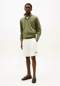 Jeune homme debout, portant un sweat-shirt olive à fermeture éclair quart, un short blanc avec logo et des mocassins marron foncé, sur fond blanc.